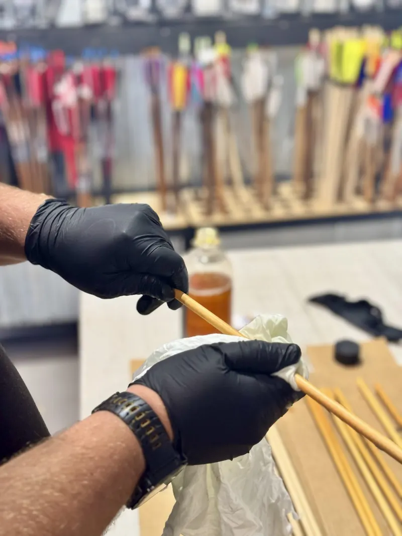 Gloved hands rub oil into a wooden arrow shaft, with finished fletched arrows racked behind