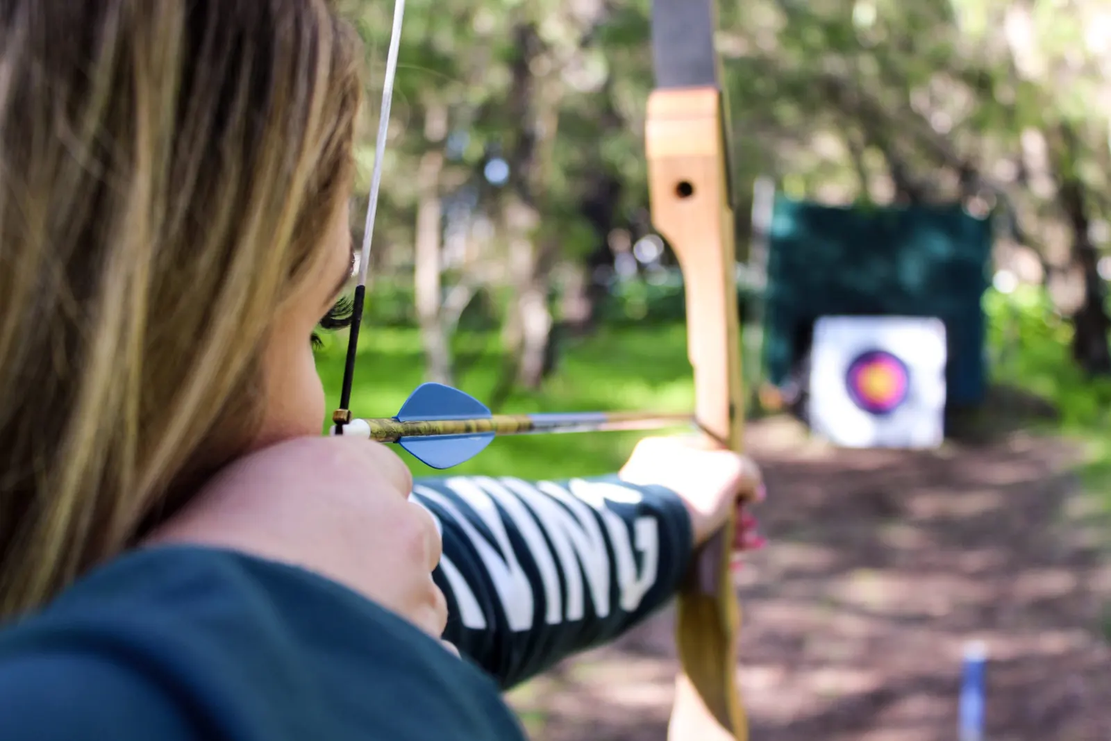 An archer draws a wooden recurve bow and aims at a target among the trees