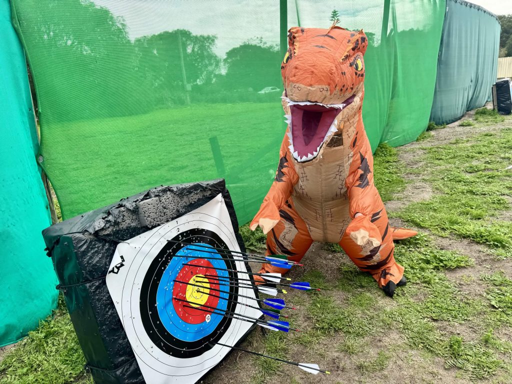 Nugget the T-rex roars with excitement beside a target full of arrows on the practice range