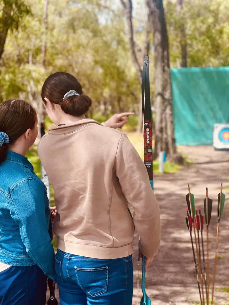 Two archers on the practice range, one pointing out the target, with arrows standing in the ground beside them