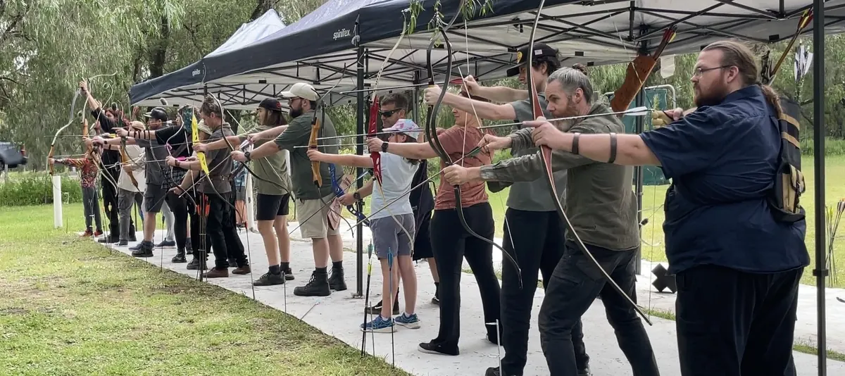 A long line of archers of all ages drawing their bows together under the shade tents