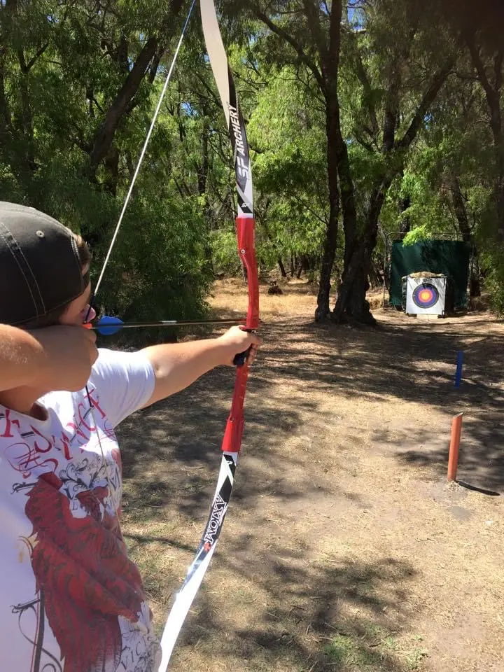 An archer in a cap draws a red and white recurve bow at a target down a shaded bush track